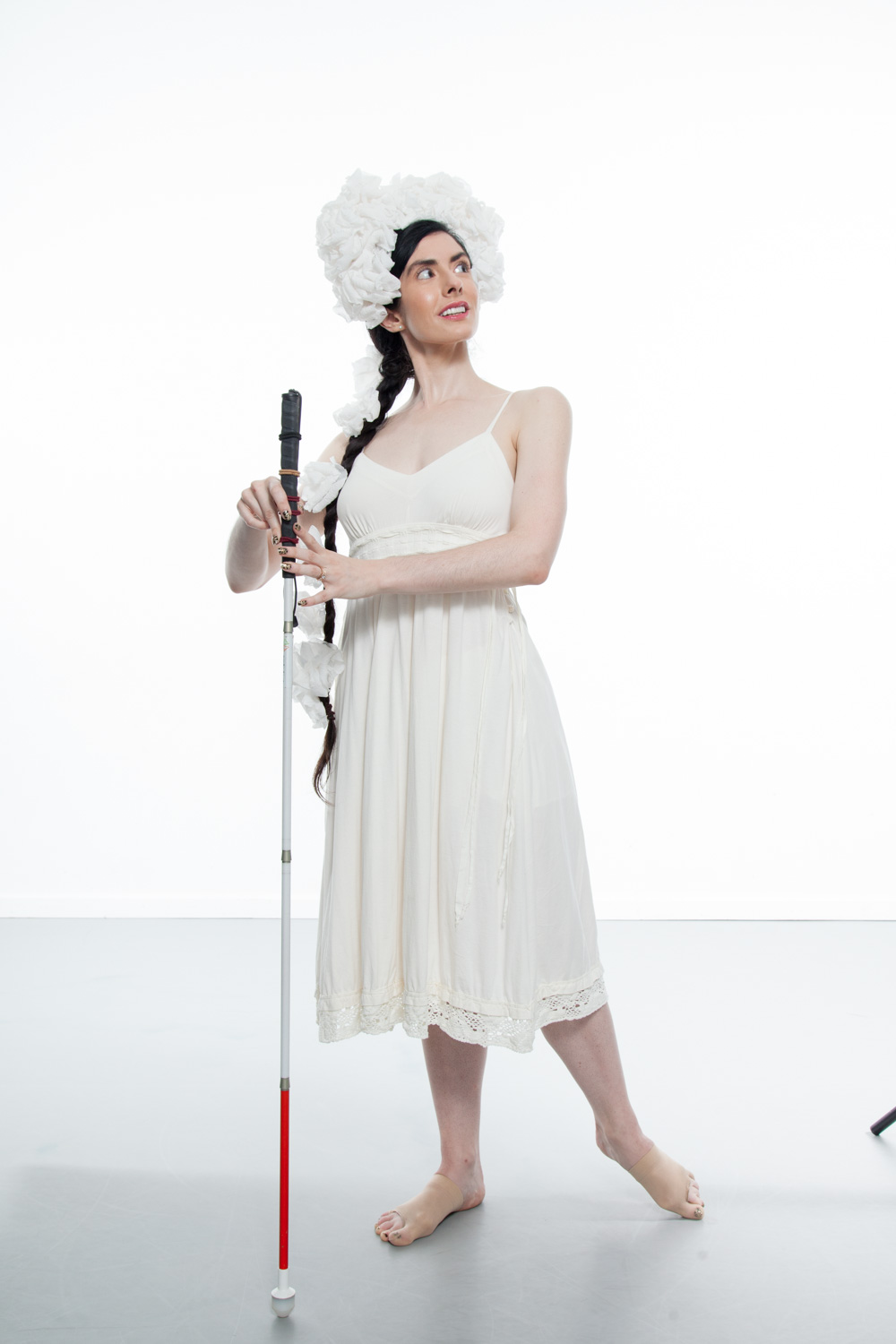 Image description: Krishna, a thin, pale skinned woman with long dark hair pulled into a braid, poses on a studio stage. She is wearing a simple white dress with her shoulders exposed and a fluffy white headdress perched on her forehead and wrapping down her braid. She looks over her left shoulder as she lightly holds a straight cane between her hands and points her left foot behind her. Photo Credit: Krishna Washburn & Micaela Mamede